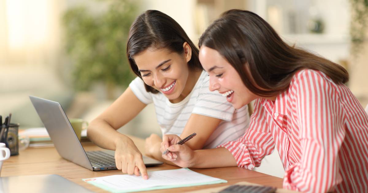 Two young women smiling while seated in front of a laptop. They are reading and signing a document on the table.