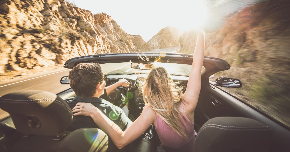 A man drives a convertible car toward the sunset in rocky terrain. A woman in the passenger seat raises her hand.