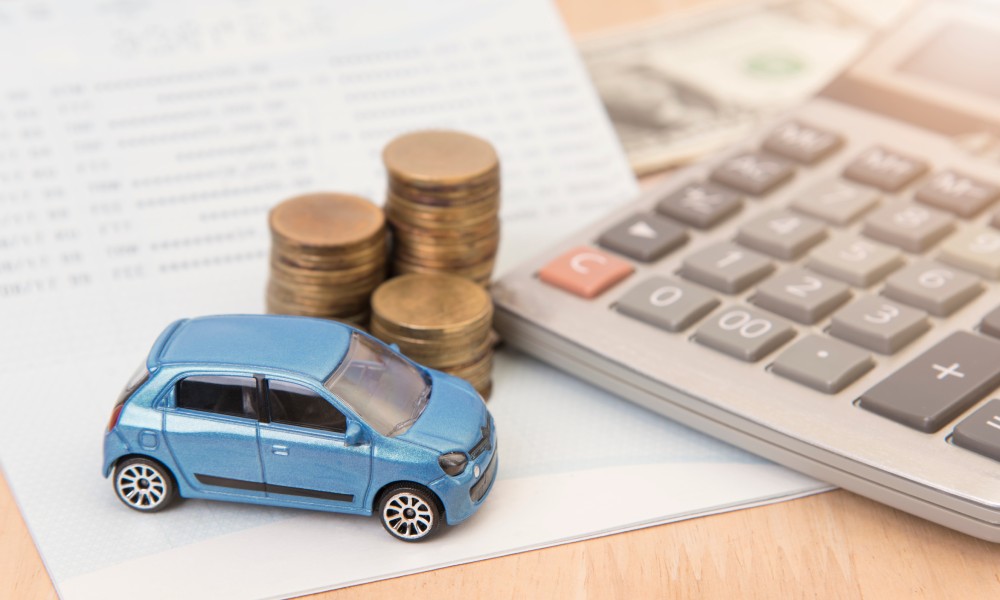 A blue toy car sits beside three stacks of coins and a calculator, placed on top of papers and dollar bills on a table.