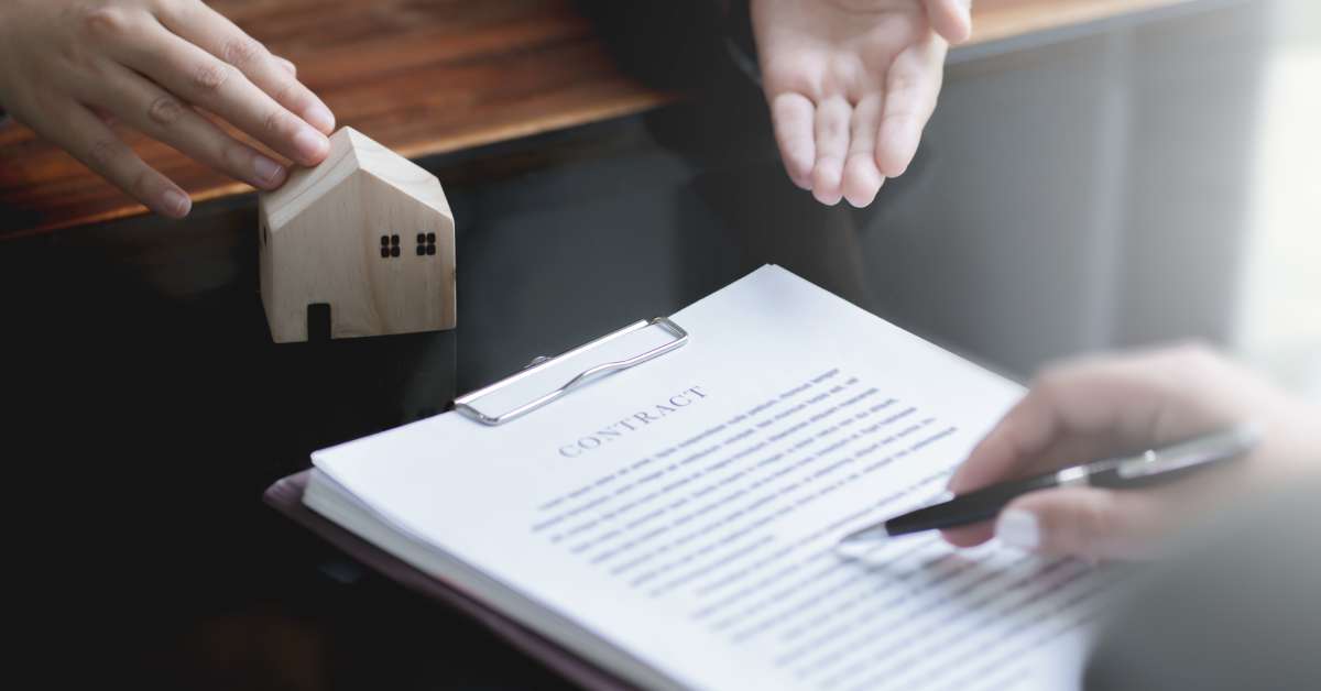 Two people review a thick contract. One man holds a pen and reads the print, while the other gestures toward a wooden house.