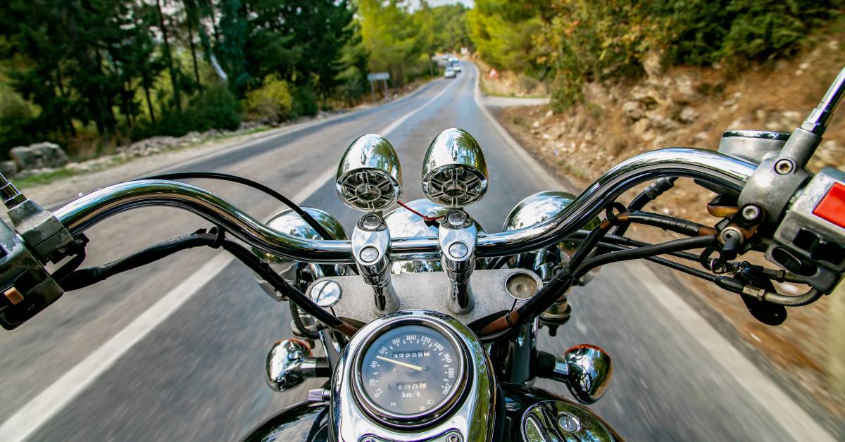 A rider's perspective of chrome motorcycle handlebars. The vehicle is headed downhill on a road with cars approaching.