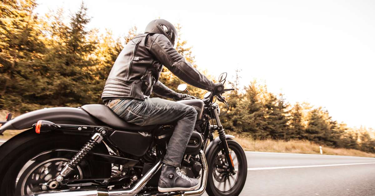 A man riding a motorcycle wearing a black helmet, leather jacket, and jeans on a road lined with trees.