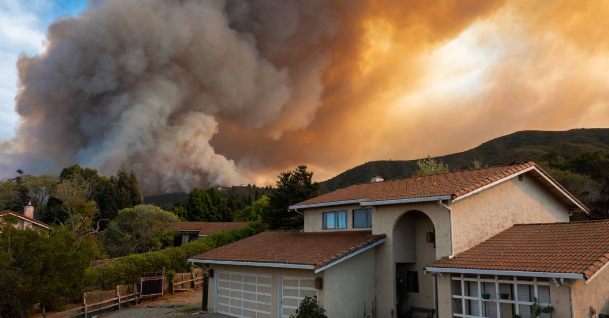 A wildfire on a forest-covered hillside with a large cloud of smoke with a Spanish-style home in the foreground.