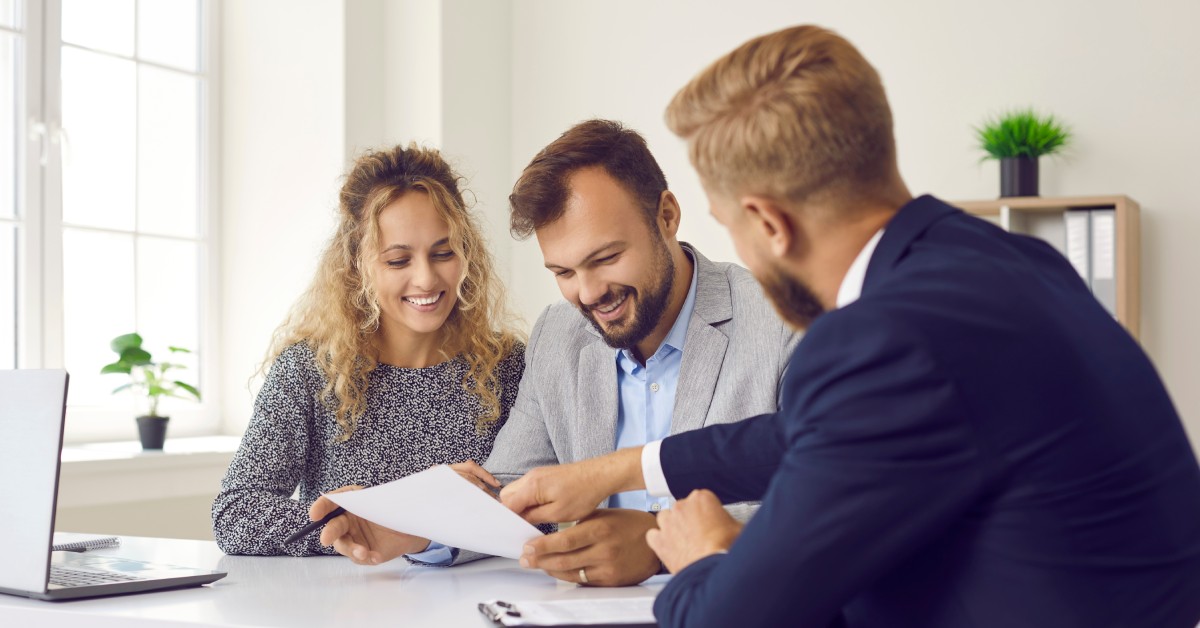 A couple speaking to a male insurance agent in his office who is gesturing toward a printed contract.