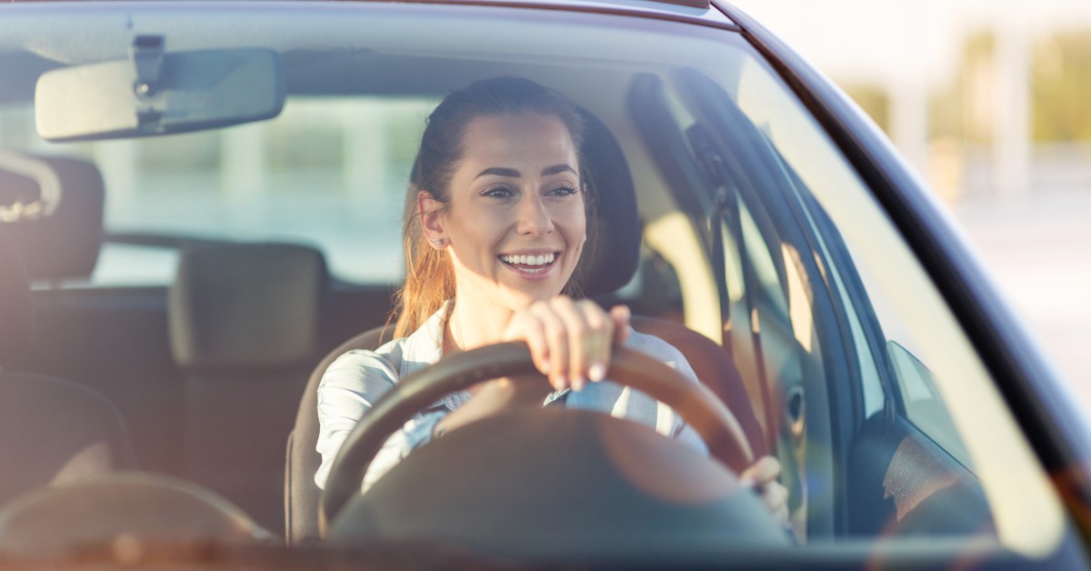 A woman in a denim shirt is driving a car during golden hour. She is smiling, and the background is blurred.