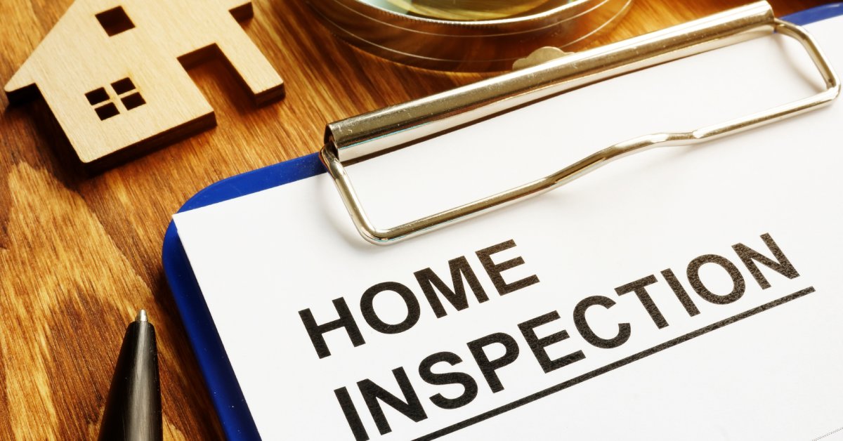 A close-up of a piece of paper reading "home inspection" on a blue clipboard near a magnifying glass and a small house.