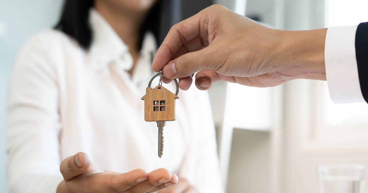 A businessman hands a key on a ring with a wooden home keychain to a woman wearing a white buttoned-down shirt.