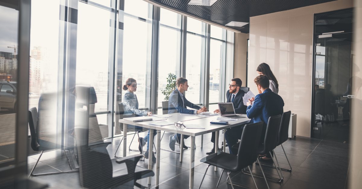 Five business professionals sit around a conference table in a meeting room with floor-to-ceiling windows.