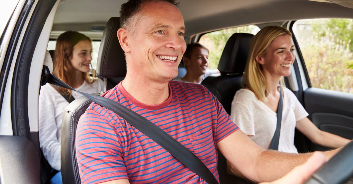 A family of four rides in a car with the father driving, the mother in the passenger seat, and two teenage children in the back.