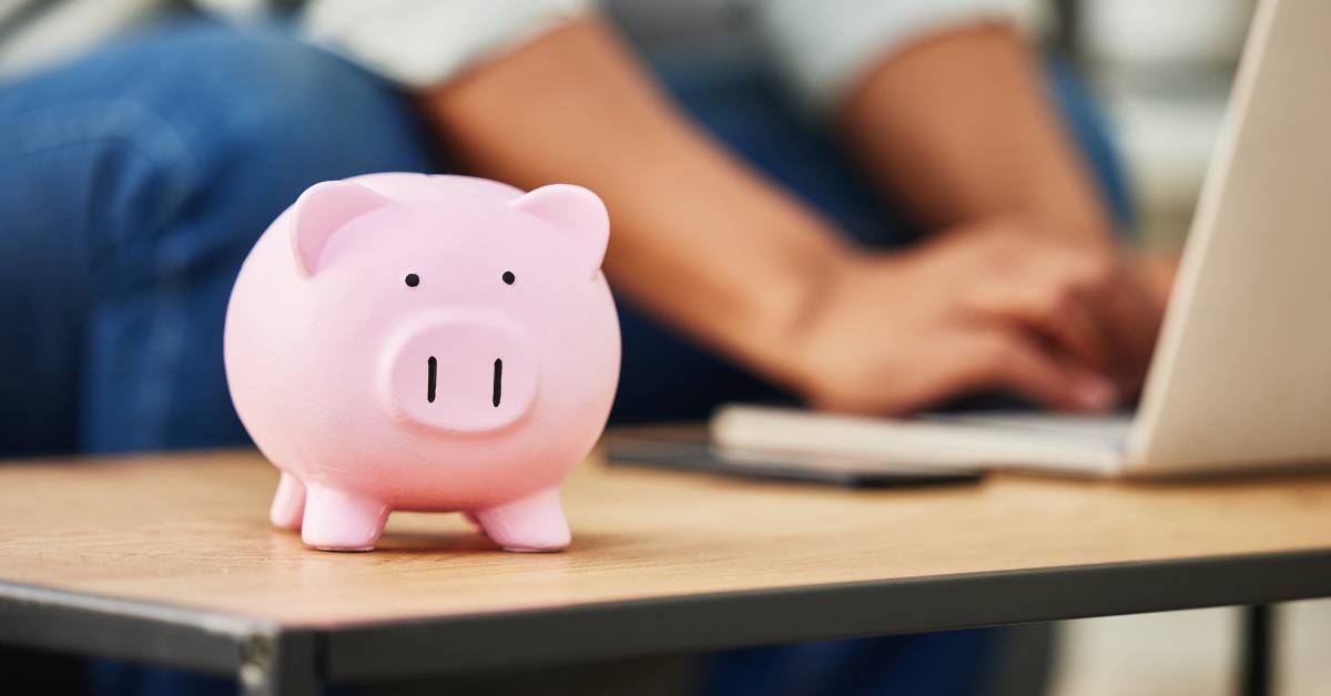 A pink piggy bank sits on a coffee table in the foreground. In the blurred background, a man types on a laptop.