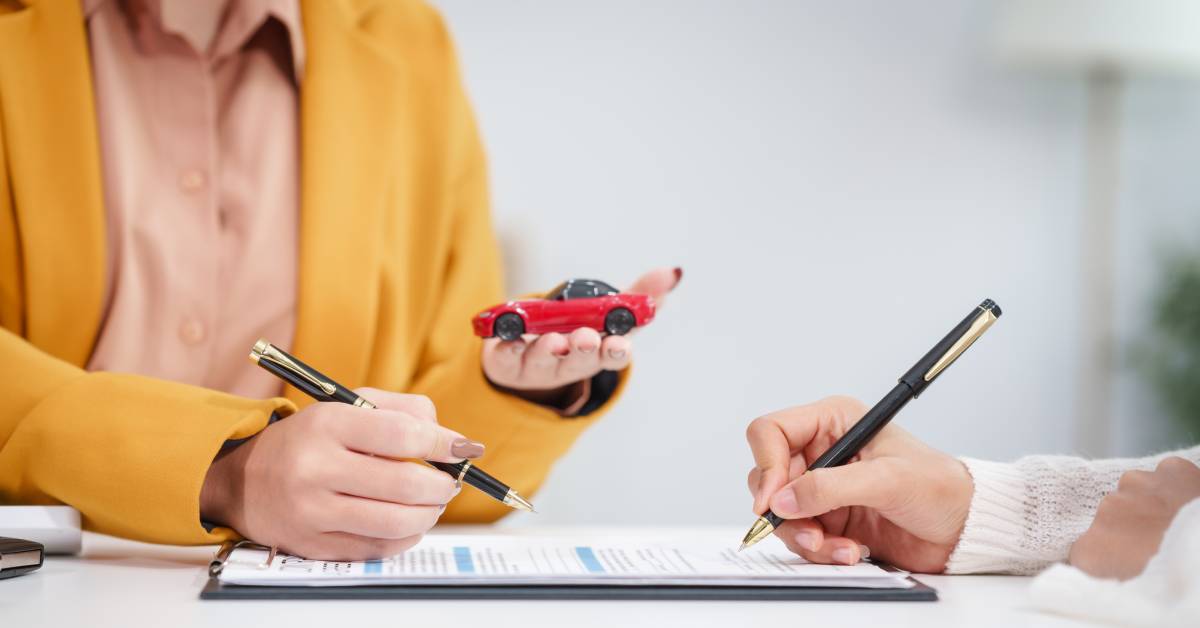 Two women hold pens, pointing to text on a contract on a black clipboard. One woman holds a toy car in her hand.