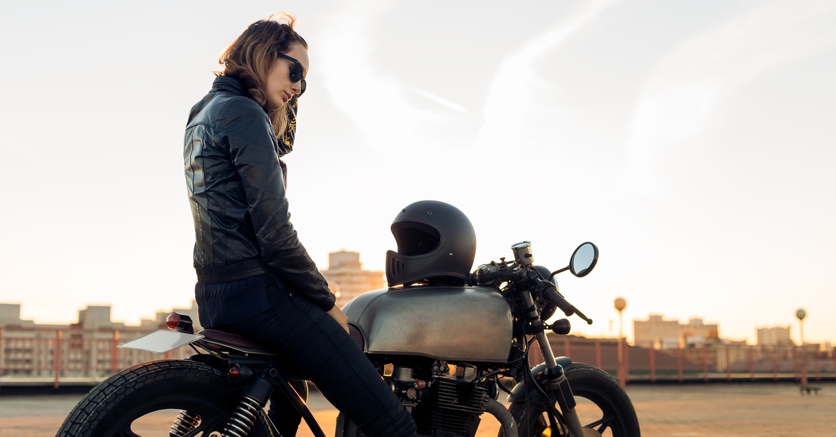 A woman wearing a black leather jacket and sunglasses sits on a motorcycle on a city roof in front of the sunset.