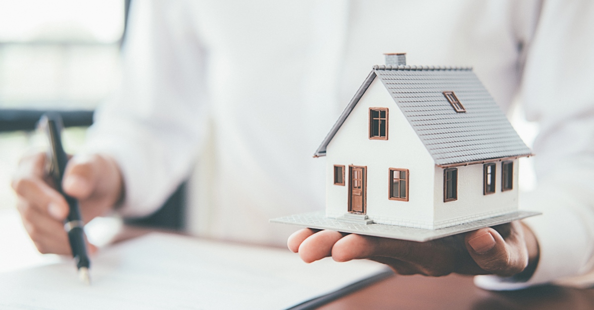 A blurry man wearing a white shirt gestures to a contract with a pen and holds an in-focus model of a home in his hand.